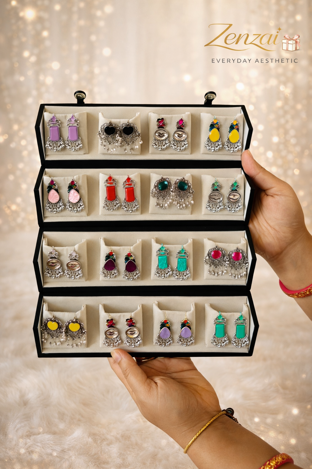 Hands holding a Zenzai 12-pair jhumka earrings gift box featuring colorful traditional silver earrings displayed in a premium velvet box with soft golden background and Zenzai logo.